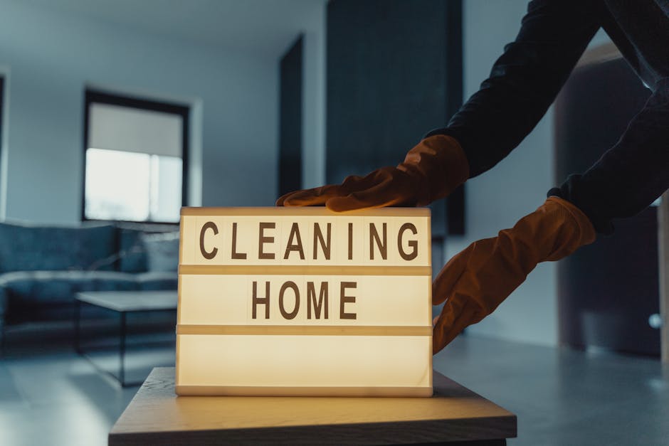A person wearing orange rubber cleaning gloves is adjusting a small illuminated sign on a wooden table that reads 'CLEANING HOME.' The background features a modern living room with a dark-colored sofa, a glass coffee table, and a window allowing natural light to illuminate the space. The scene emphasizes domestic surface cleaning and deep cleaning practices, aligning with end of tenancy cleaning tips in Kingston KT1, Kingston upon Thames. Kingston upon Thames Cleaners specializes in hygienic surface sanitisation, ensuring carpets, floors, and furniture are thoroughly cleaned and maintained for a renewed, spotless home environment.
