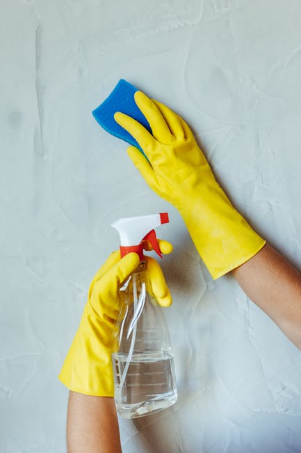Close-up of a person’s hands wearing yellow rubber cleaning gloves, engaged in surface cleaning. One hand is holding a blue sponge, wiping a light-colored, smooth wall or surface, while the other hand is holding a clear spray bottle filled with cleaning solution. The background shows a clean, minimally textured surface, indicative of a domestic or commercial environment. The image emphasizes surface sanitation, deep cleaning, and hygiene practices carried out by Kingston upon Thames Cleaners as part of end of tenancy cleaning, demonstrating attention to detail and effective cleaning tools for maintained cleanliness in residential or commercial spaces.