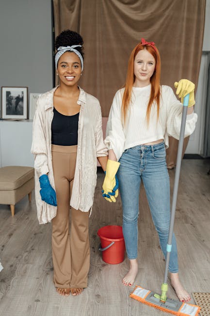 A person wearing orange rubber cleaning gloves is adjusting a small illuminated sign on a wooden table that reads 'CLEANING HOME.' The background features a modern living room with a dark-colored sofa, a glass coffee table, and a window allowing natural light to illuminate the space. The scene emphasizes domestic surface cleaning and deep cleaning practices, aligning with end of tenancy cleaning tips in Kingston KT1, Kingston upon Thames. Kingston upon Thames Cleaners specializes in hygienic surface sanitisation, ensuring carpets, floors, and furniture are thoroughly cleaned and maintained for a renewed, spotless home environment.