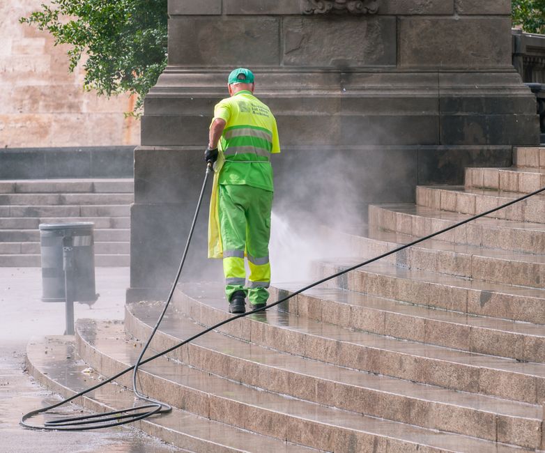 A commercial cleaning trolley and waste bin situated against a beige tiled wall in a commercial or industrial setting. The trolley is filled with cleaning tools such as mops, brushes, spray bottles, a
