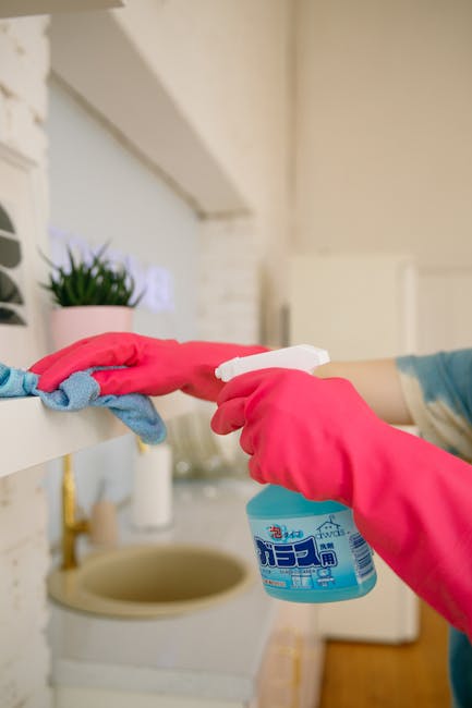 Close-up of a person wearing pink rubber gloves using a spray bottle of cleaning solution to disinfect a white shelf in a kitchen. A small potted green plant sits on the shelf, which is above a beige