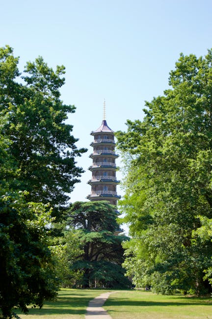 An outdoor scene featuring a tall, traditional multi-tiered pagoda surrounded by lush, green trees and a well-maintained grassy pathway leading towards it. The pagoda has a grey slate roof with multip